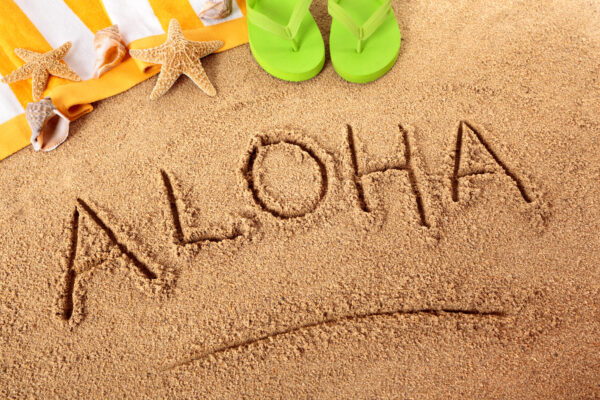 The word Aloha written on a sandy beach, with flip flops, towel, starfish and seashells in the background.  Sharp focus on Aloha and distant objects slightly blurred.  Studio shot - directional lighting and warm color are intentional.